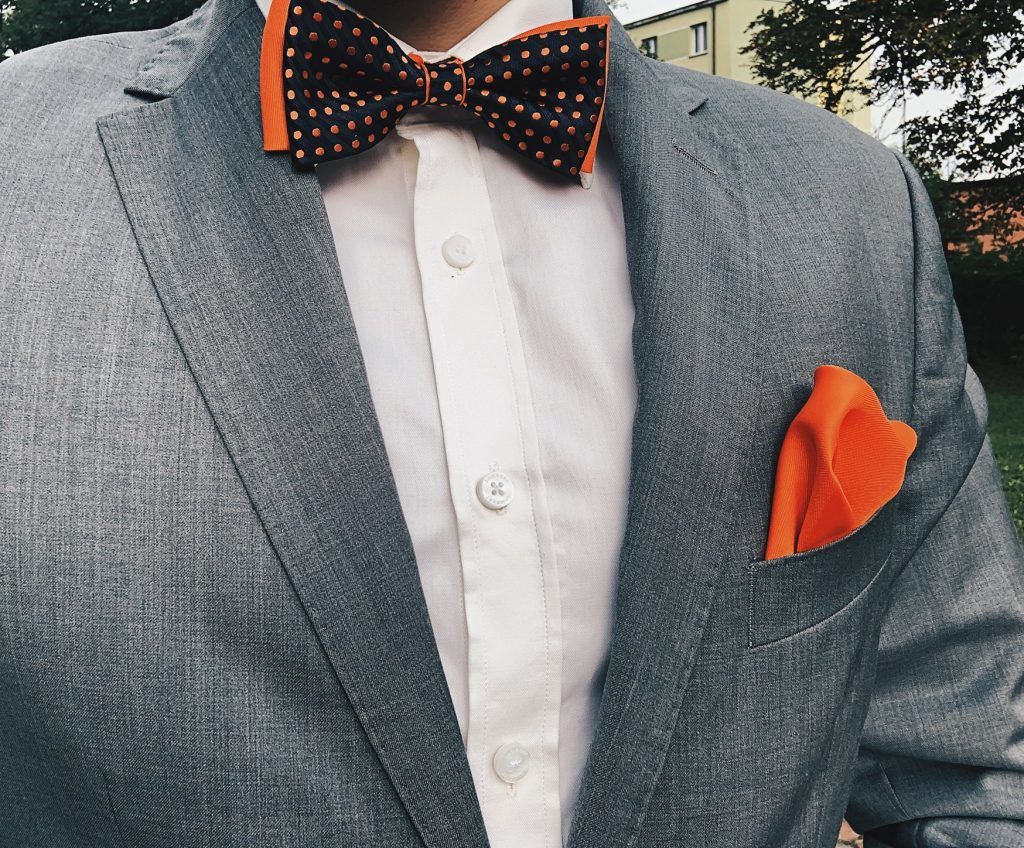 Szary garnitur z dodatkami: pomarańczowa muszka oraz pomarańczowa poszetka. Jak ubrać się na wesele?